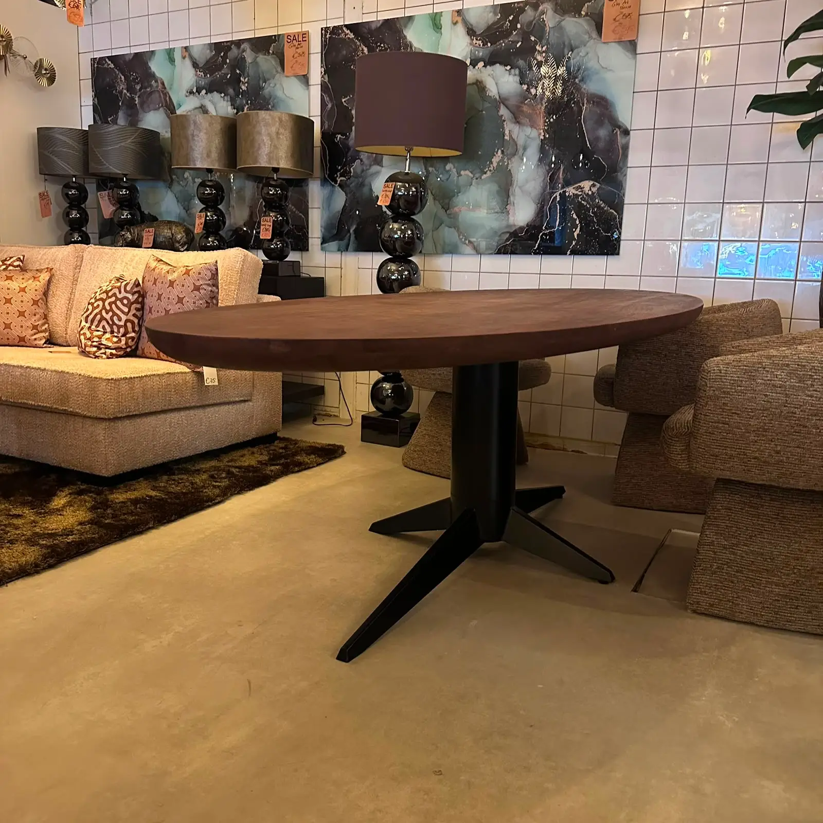 Modern living room showroom: round wooden table on a black pedestal, beige sofa with patterned cushions, marble-blue wall art, and stacked-glass lamps along a tiled wall.