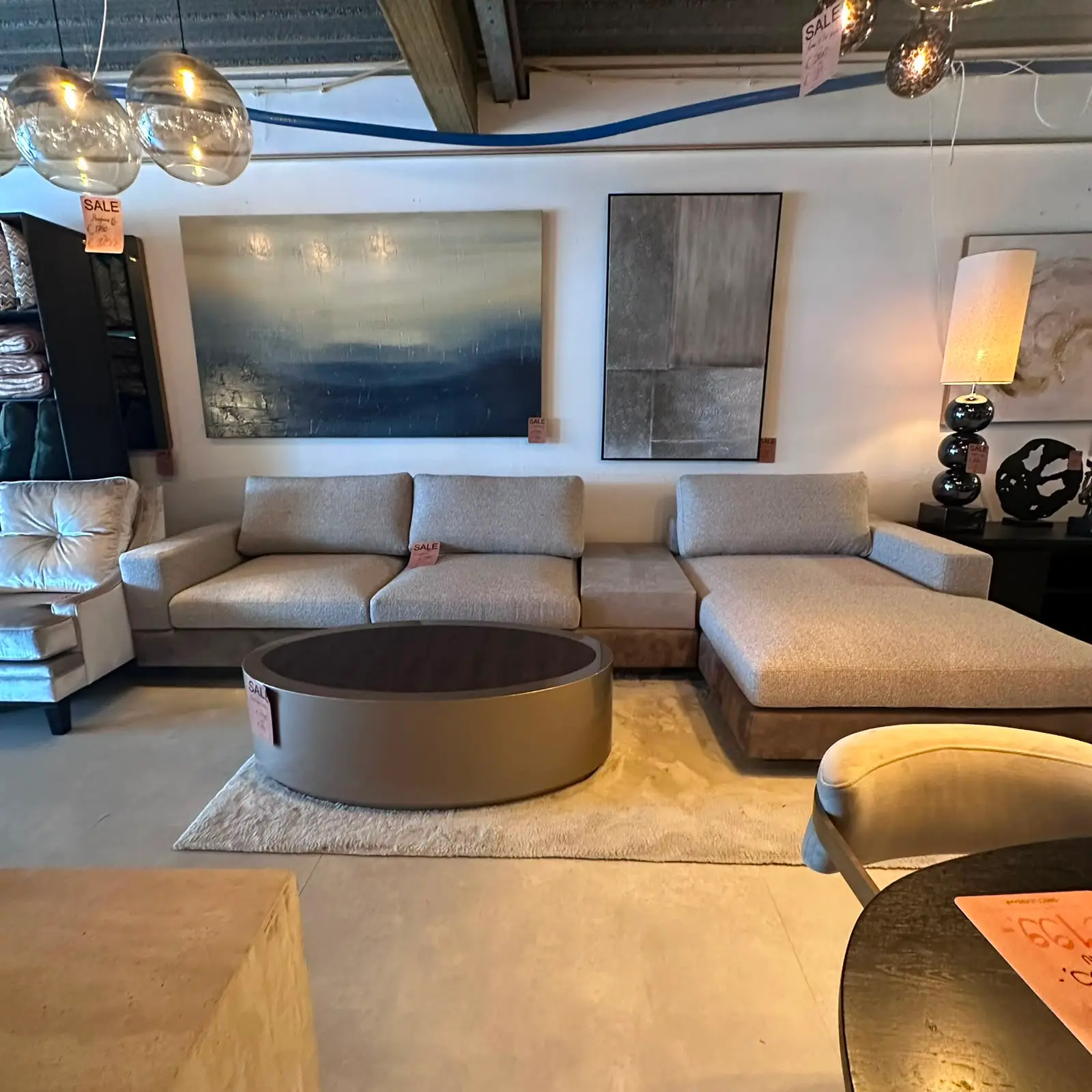Modern showroom living room with a light gray sectional sofa, ottoman, and round coffee table on a beige rug (sale tag visible).