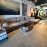 Gray sectional sofa on display in a furniture showroom, with a round metal coffee table and sale tag nearby