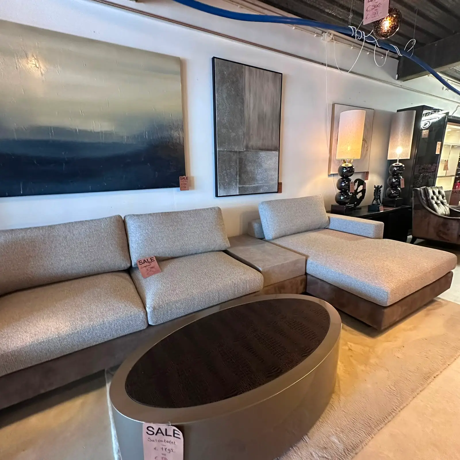 Modern showroom with a light gray sectional sofa and a black circular coffee table, beige carpet, and abstract wall art above the sofa; sale tag attached to cushions.