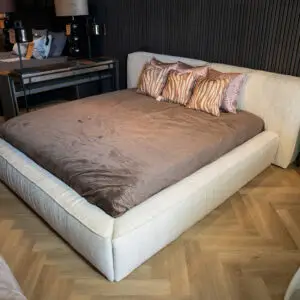 Wide view of a large beige upholstered bed with brown quilt and zebra-pattern pillows in a modern bedroom; wood herringbone floor and dark paneled wall in the background