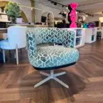 Two green patterned swivel chairs on a wooden floor in a showroom, with a large pink Gucci balloon sculpture in the background and decorative items nearby.