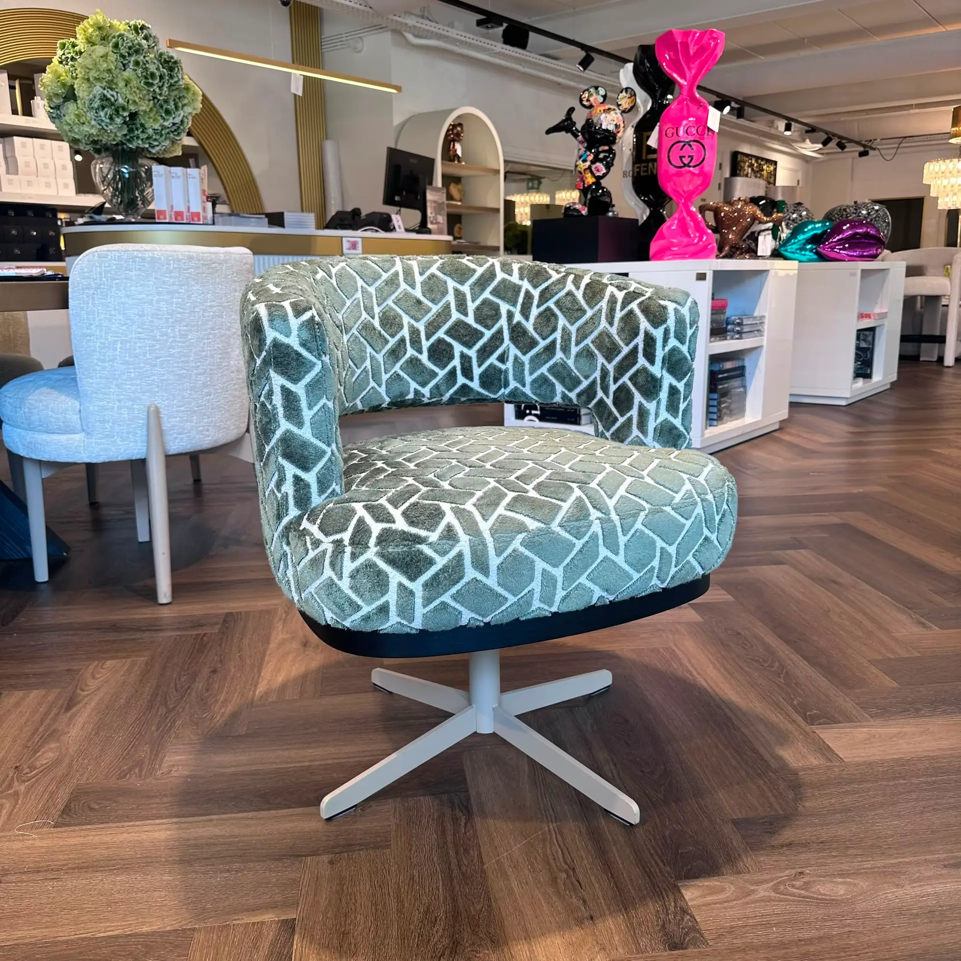 Two green patterned swivel chairs on a wooden floor in a showroom, with a large pink Gucci balloon sculpture in the background and decorative items nearby.