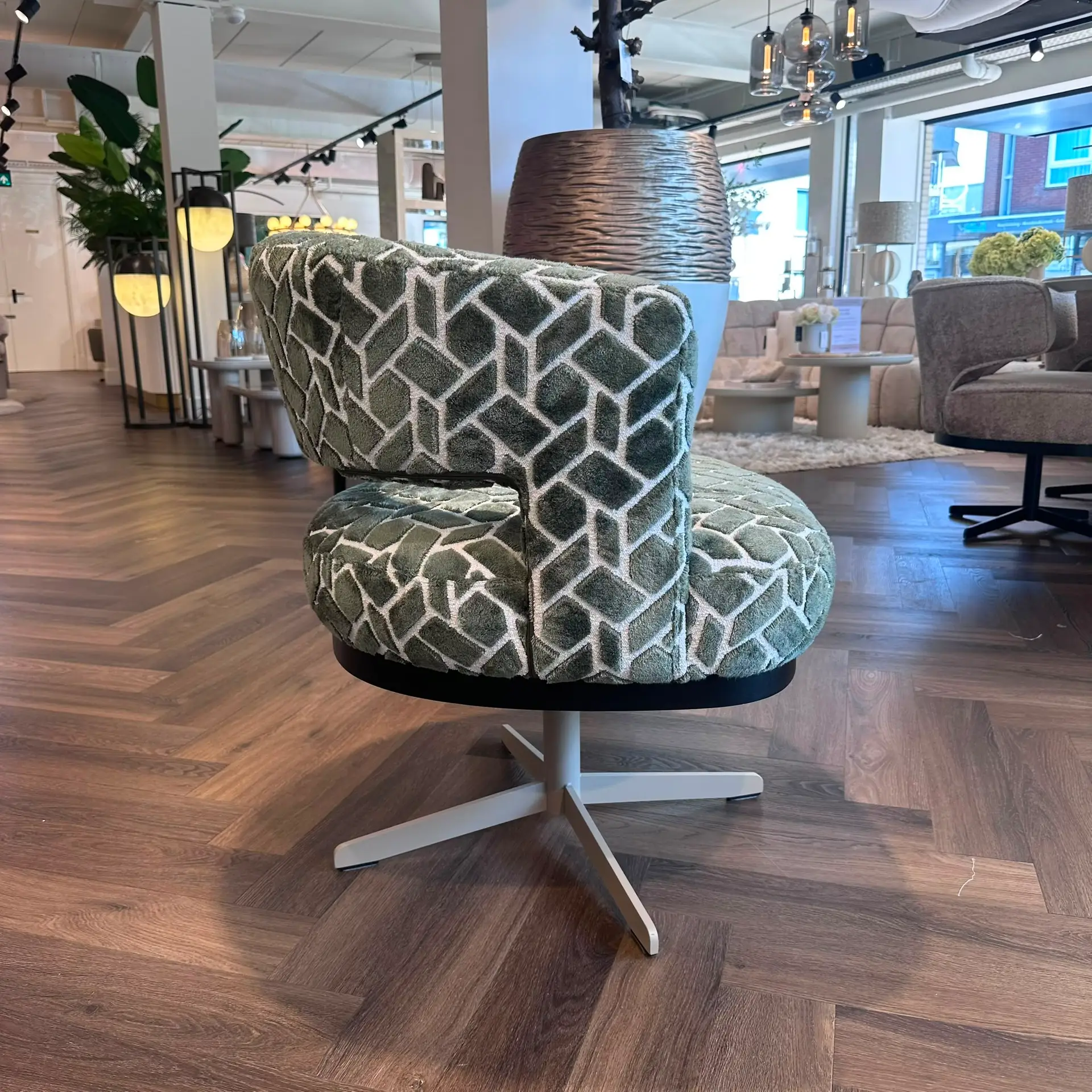 Close-up of a green geometric-patterned upholstered chair in a modern furniture store, with wooden flooring and contemporary lighting in the background.