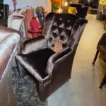 Dark gray velvet tufted armchairs in a furniture shop with sale tags visible in the foreground and colorful decor in the background.