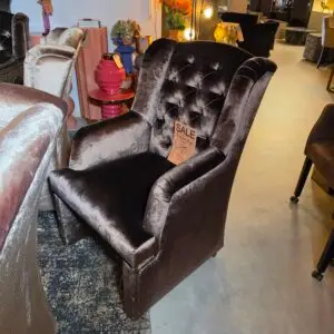 Dark gray velvet tufted armchairs in a furniture shop with sale tags visible in the foreground and colorful decor in the background.