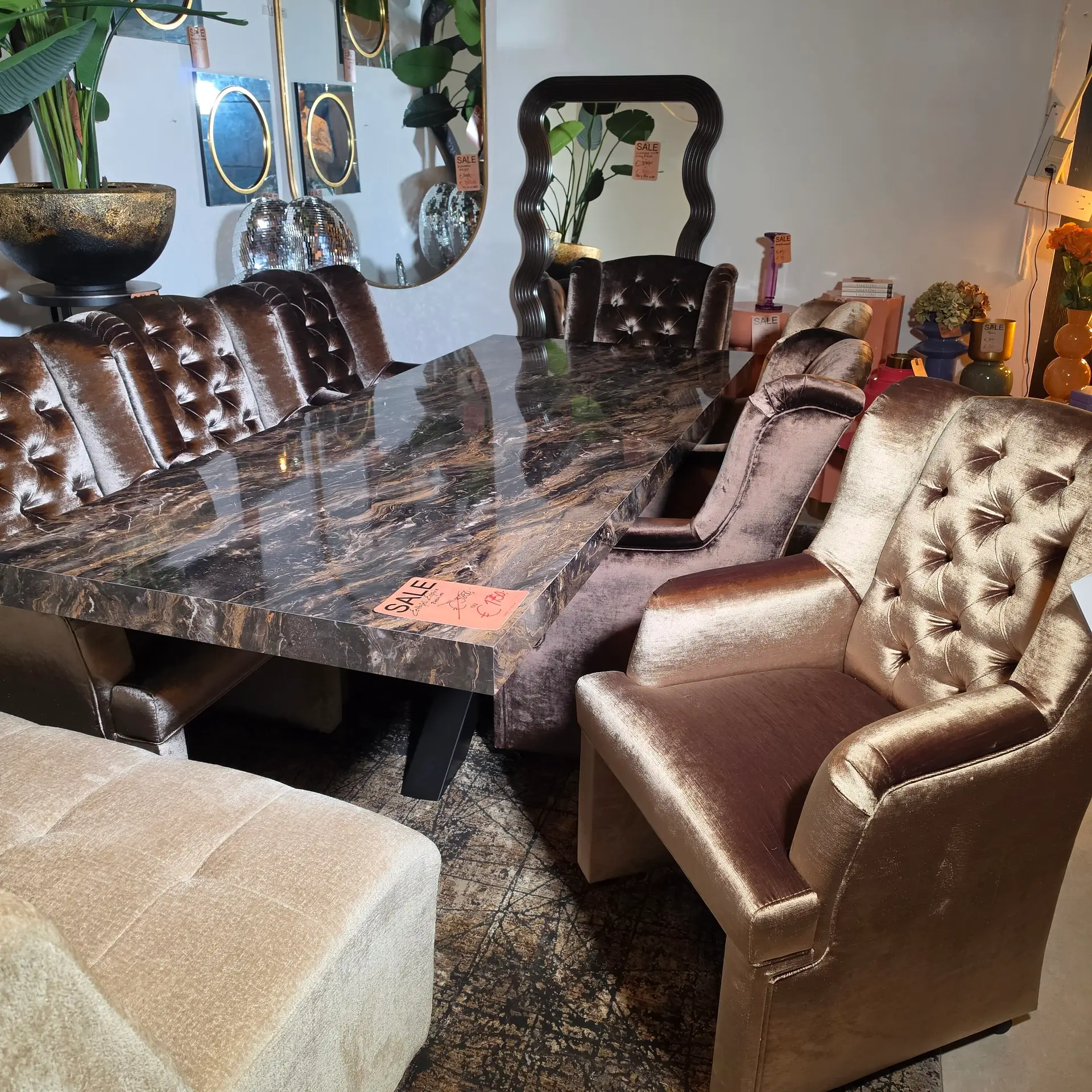 Boutique showroom with tufted brown velvet chairs around a dark marble table, sale tags visible on items and decorative mirrors in the background.