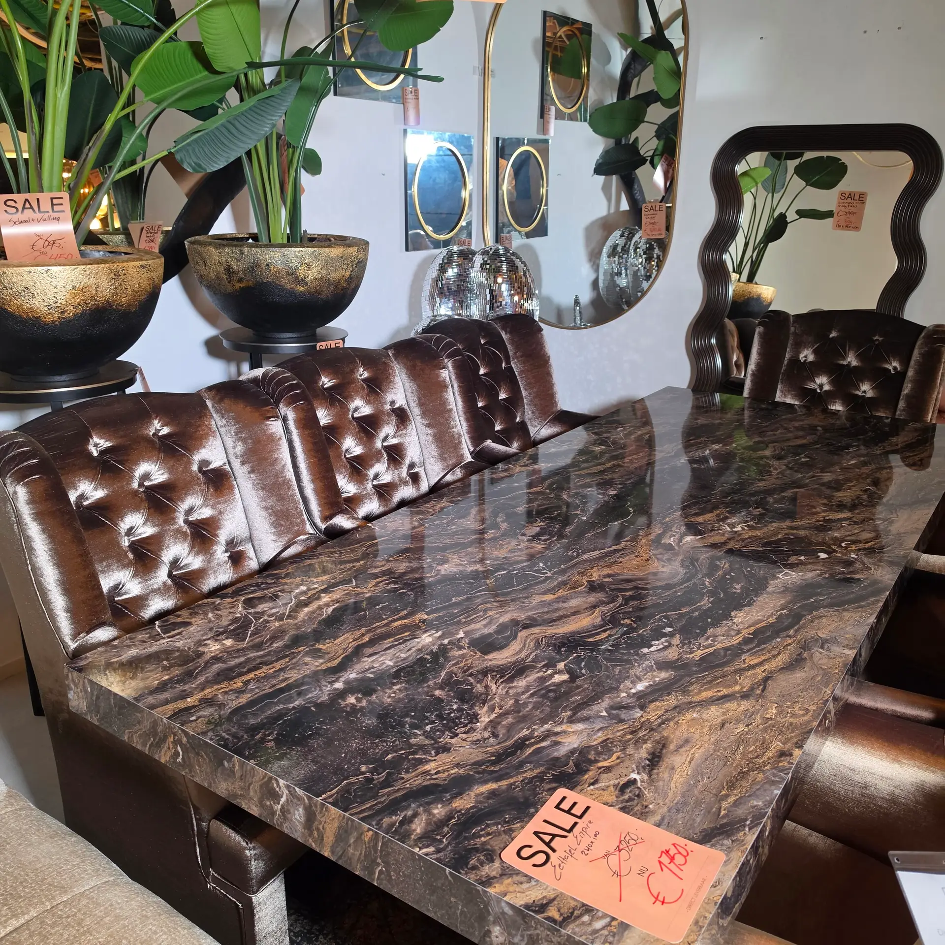 Showroom display with brown tufted leather seating, marble counters, and green potted plants with sale tags nearby.
