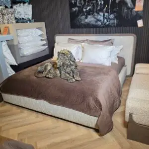 Bedroom showroom with brown quilted bedspread, cream headboard, and white pillows; a lion sculpture rests on the duvet.