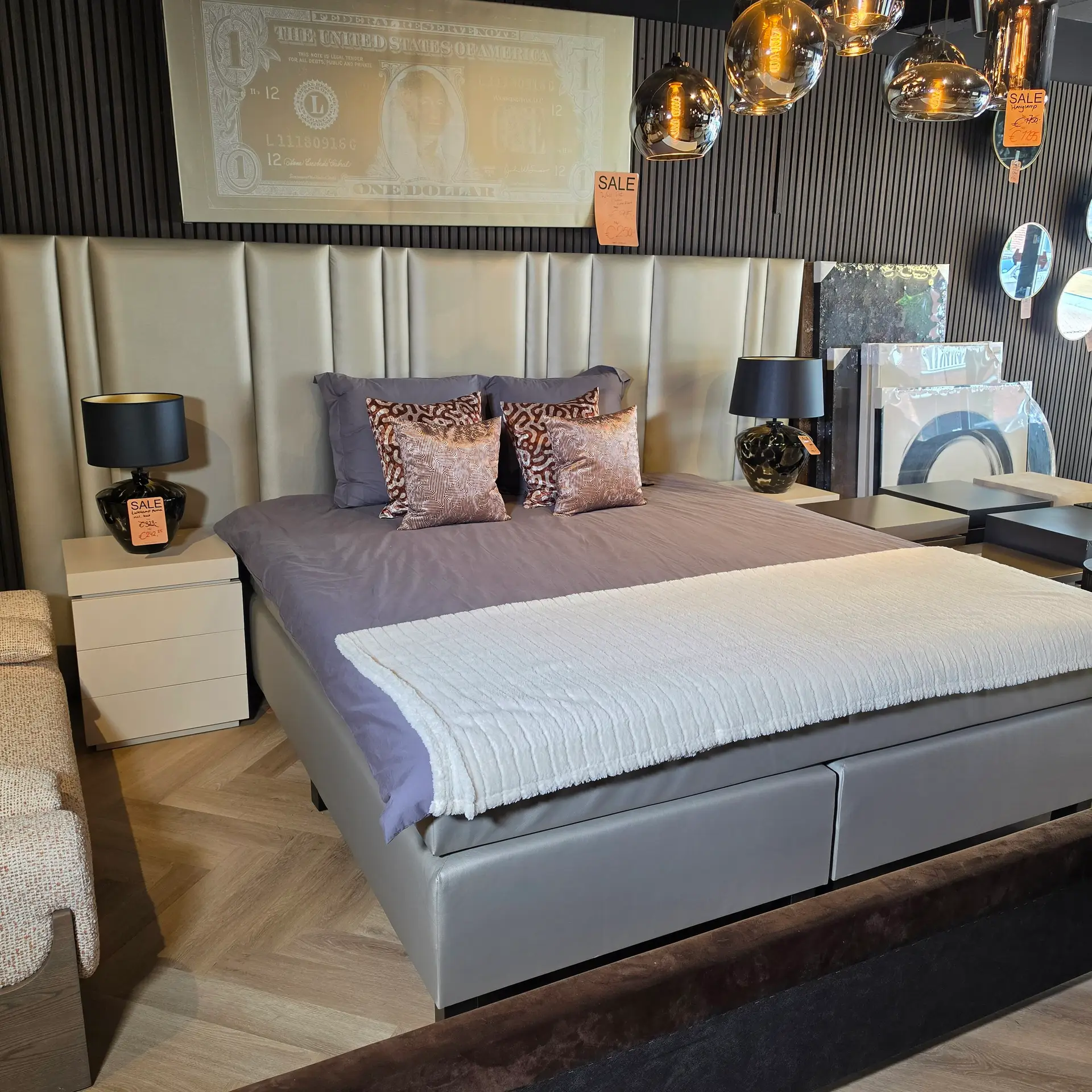 Bedroom display: large grey upholstered bed with matching nightstands, grey bedding, and patterned throw pillows with sale tags visible.