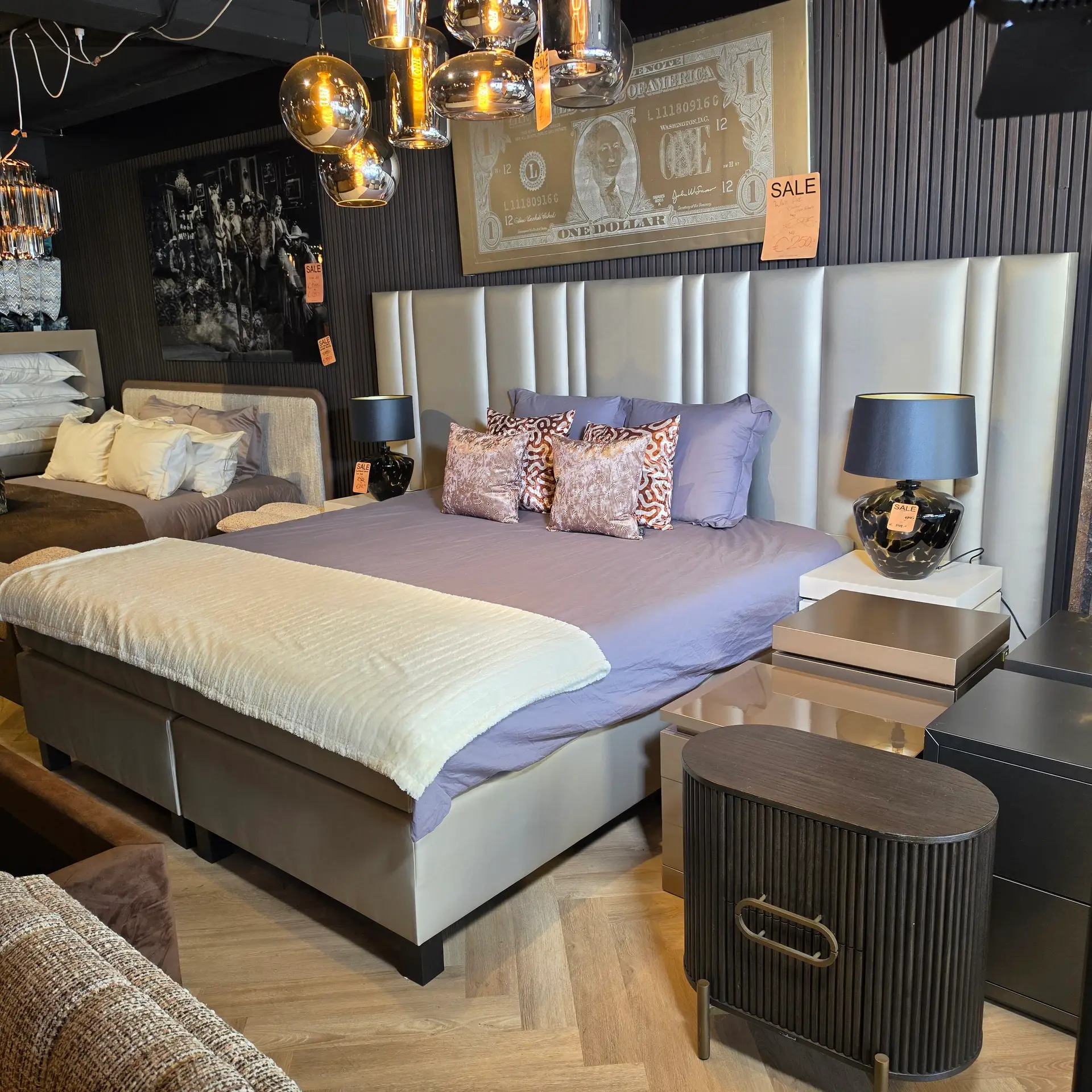 Showroom bedroom with two connected beds in lavender bedding, white throws, and glossy black nightstands with sale tags on decor.