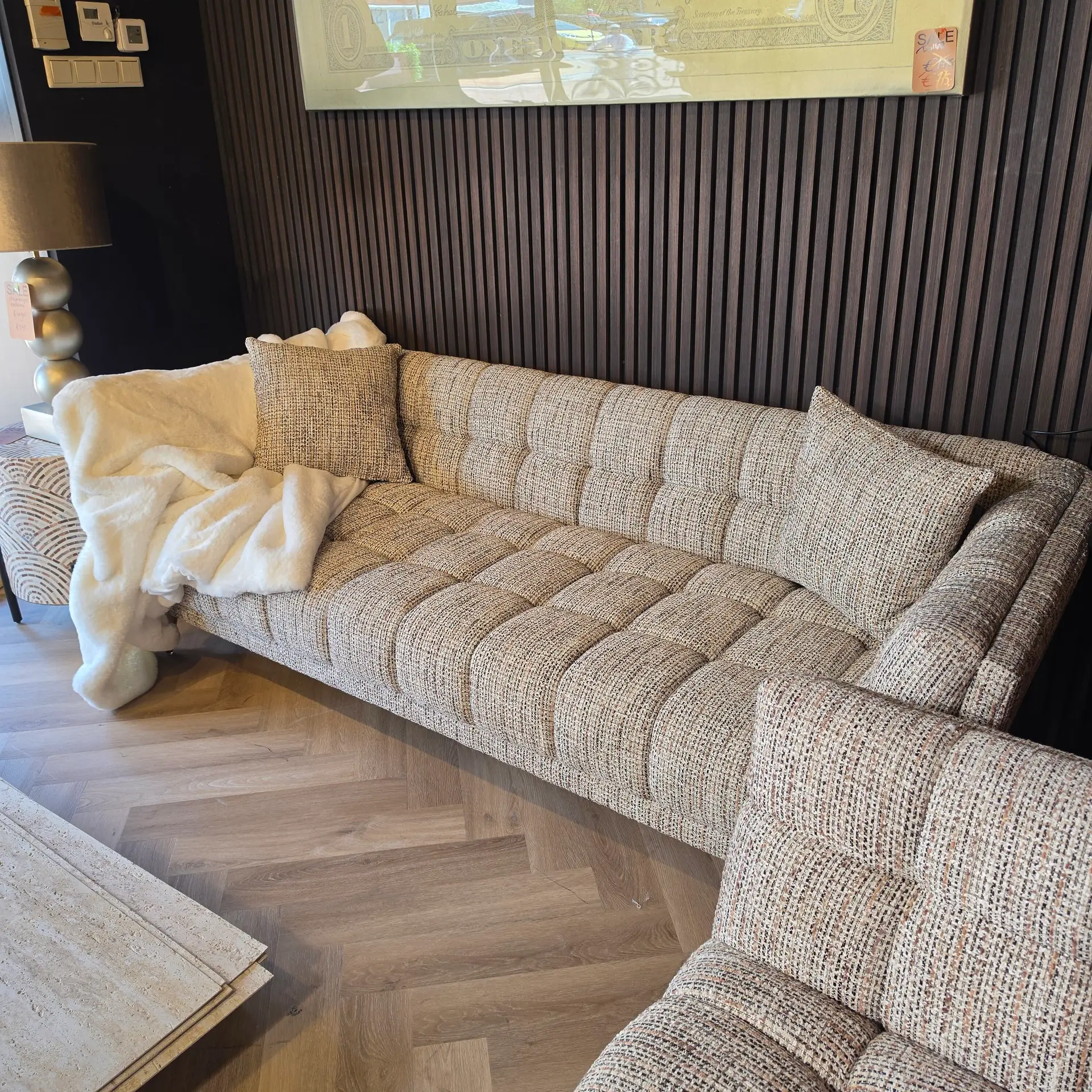 Cozy living room display with a beige-brown striped sofa, cream blanket, and a textured wall with vertical panels; sample floorboards on the left and a lamp nearby.