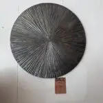 Round textured metal wall sculpture with radiating spokes in dark bronze, mounted on a pale wall; a sale tag hangs below.