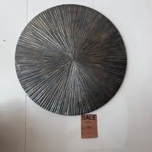 Round textured metal wall sculpture with radiating spokes in dark bronze, mounted on a pale wall; a sale tag hangs below.