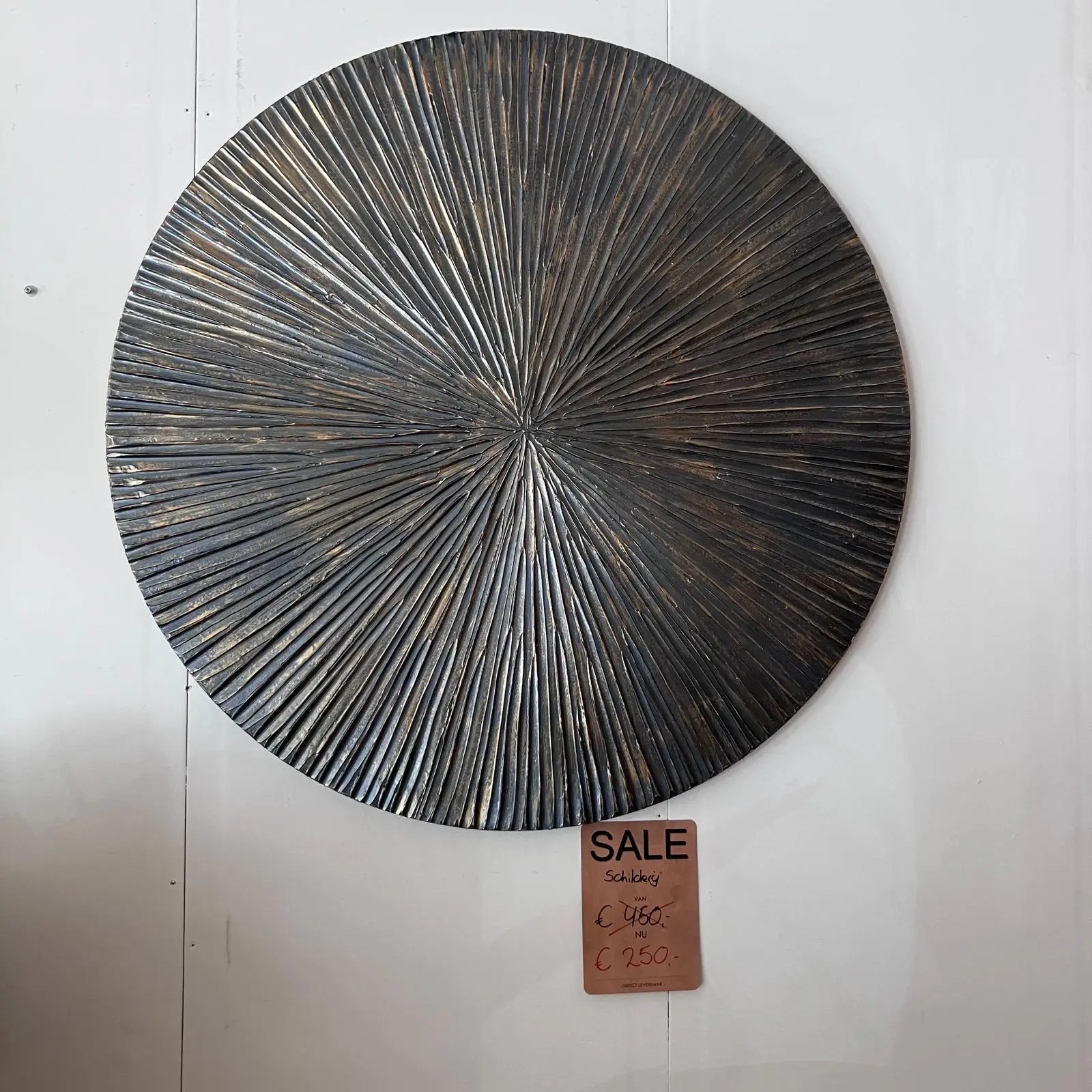 Round textured metal wall sculpture with radiating spokes in dark bronze, mounted on a pale wall; a sale tag hangs below.