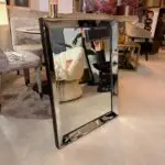 Full-length rectangular mirror with beveled edges on a black stand in a furniture showroom, reflecting beige chairs and pink flowers; sale tag visible.