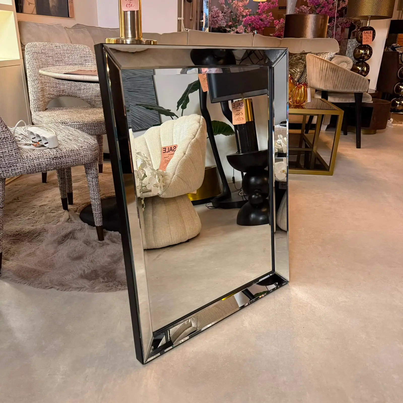 Full-length rectangular mirror with beveled edges on a black stand in a furniture showroom, reflecting beige chairs and pink flowers; sale tag visible.
